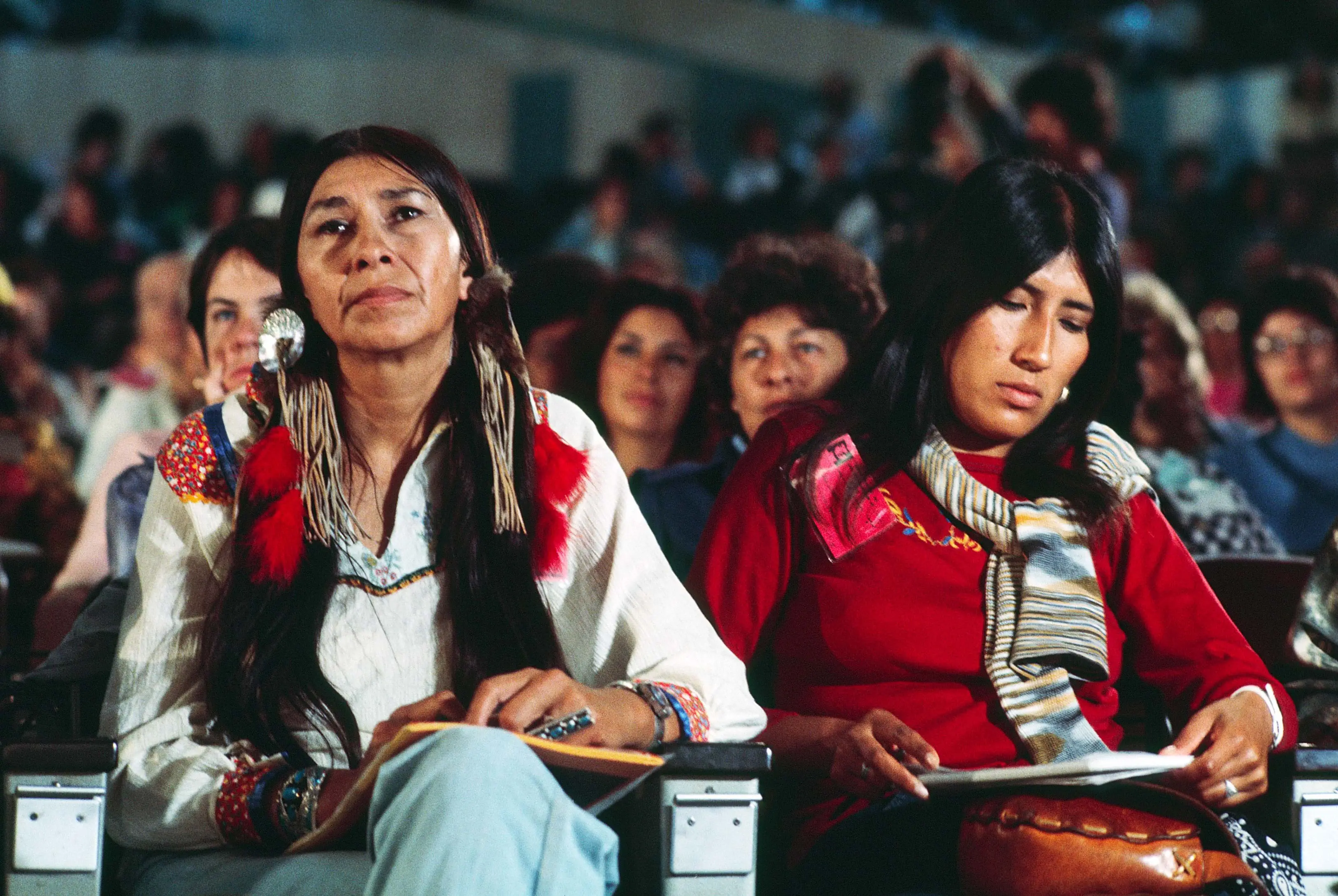 Two indigenous women sit in the front row of an audience. 