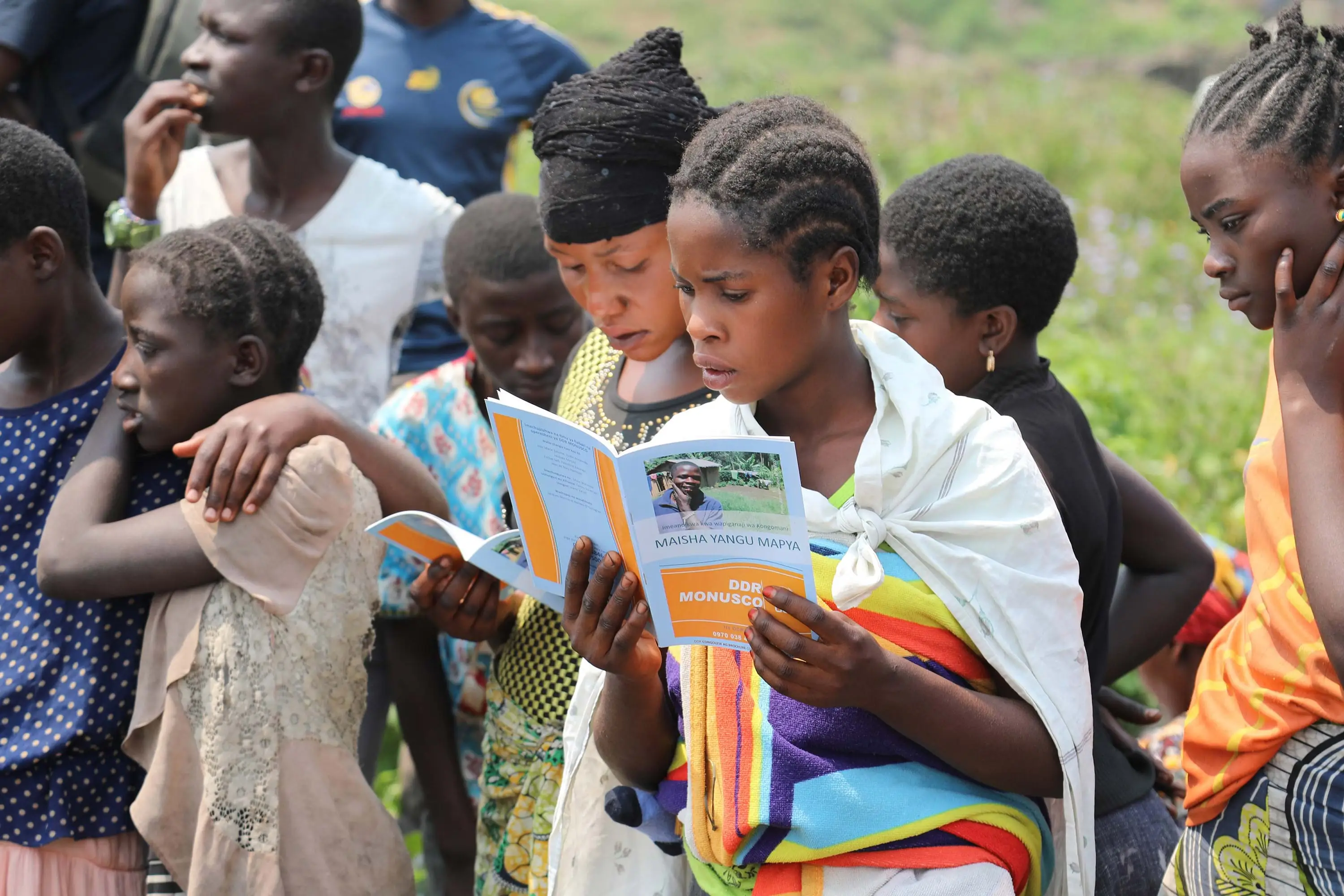 Black children stand outside. One of the children is reading a book.