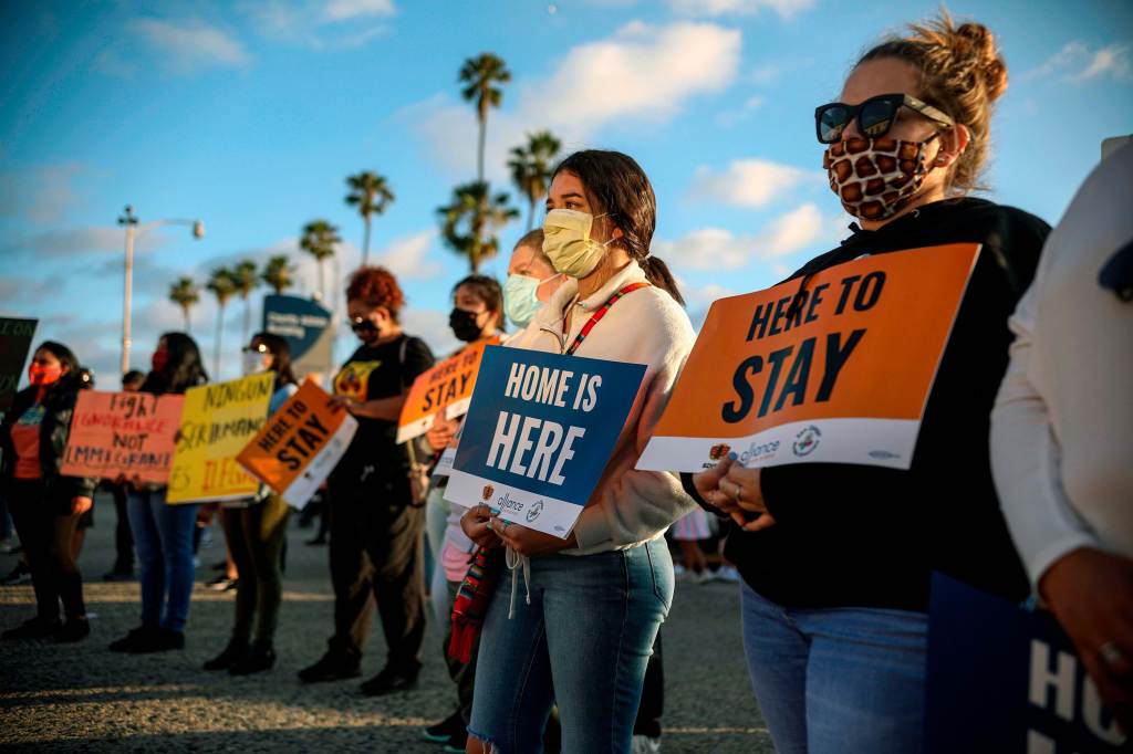 People wearing protective masks while holding signs during a rally with palm trees and blue skies behind them.