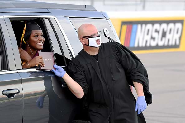 A high school principal wearing a protective  mask and gloves presents a graduate with her diploma through the car window.