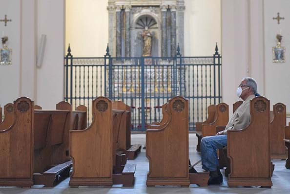 A lone man wearing a protective mask sits on a pew in an empty church.