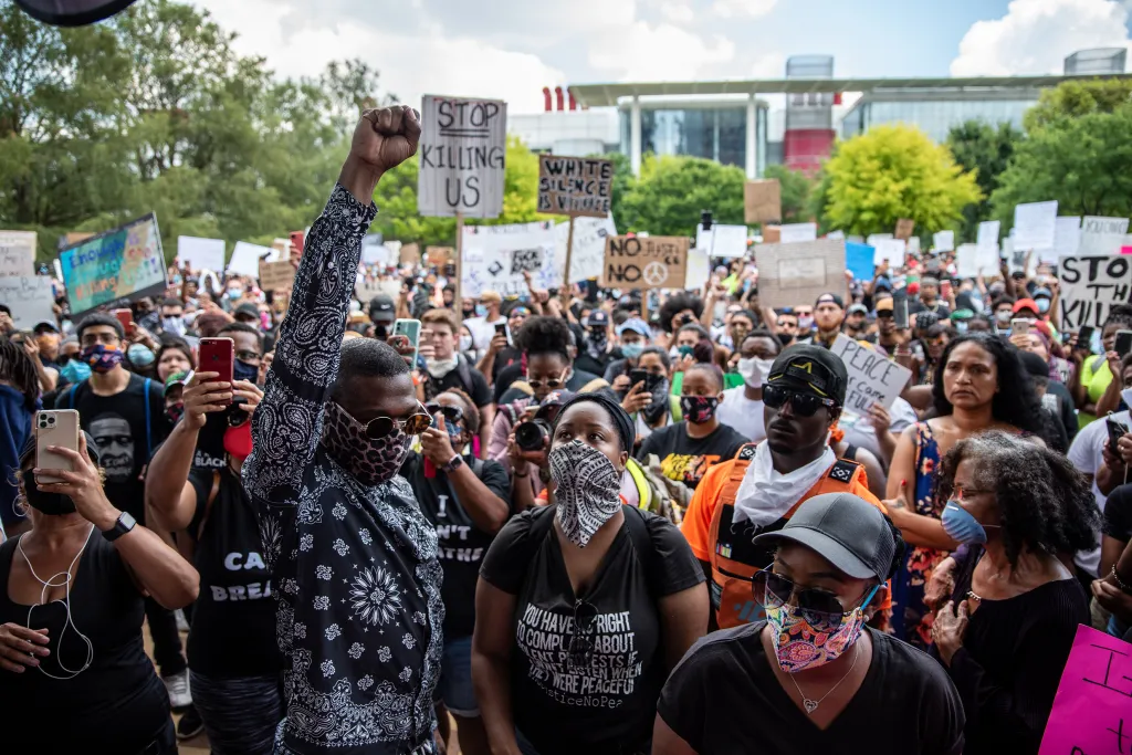 A Black man wearing a mask raises his fist among a diverse crowd of protesters, some carrying signs.