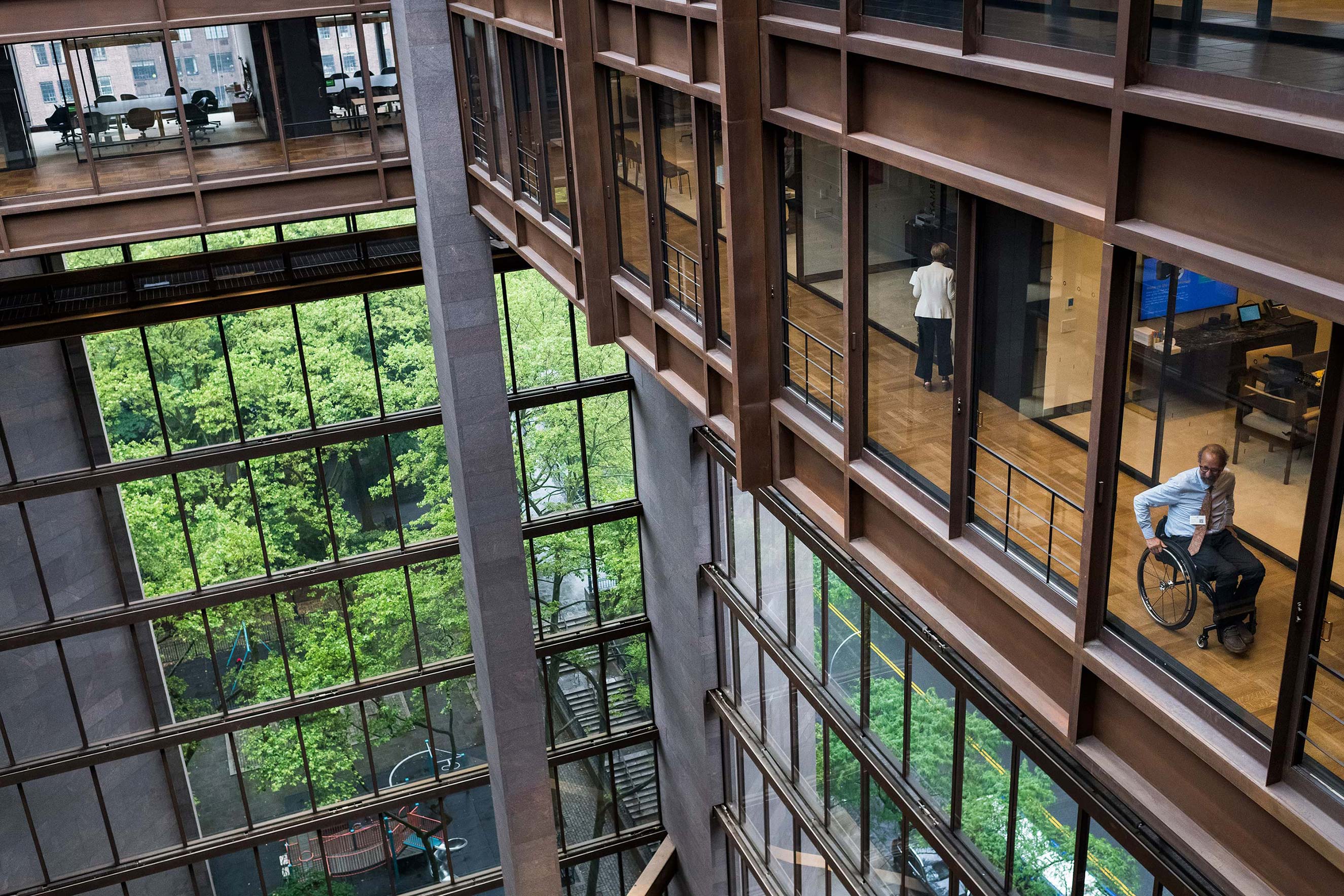 From a vantage point above the Ford Foundation’s glass and steel atrium, a man in a wheelchair looks out the window as he moves down a hallway.