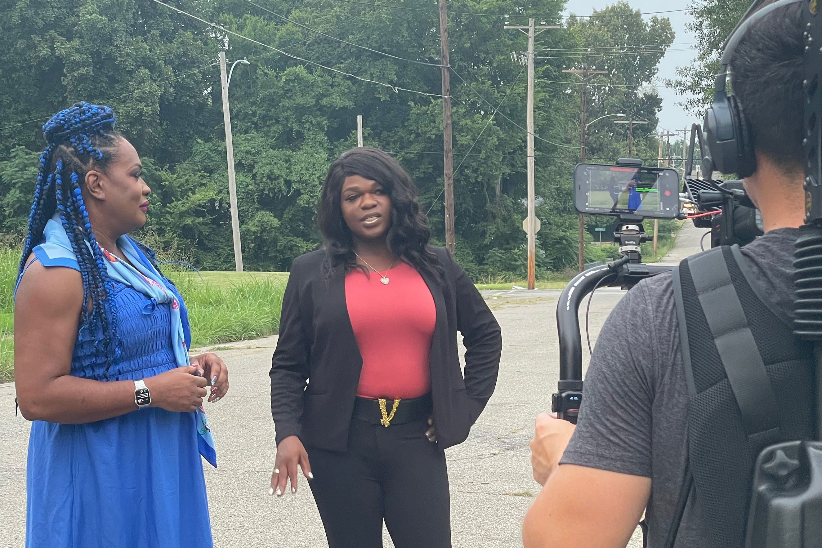 Two Black transgender women are interviewed outdoors by a videographer on the right. The woman on the left has blue braids; the woman on the right wears a black blazer over a red top. Focus: TransLash Media, LGBTQ+ rights.