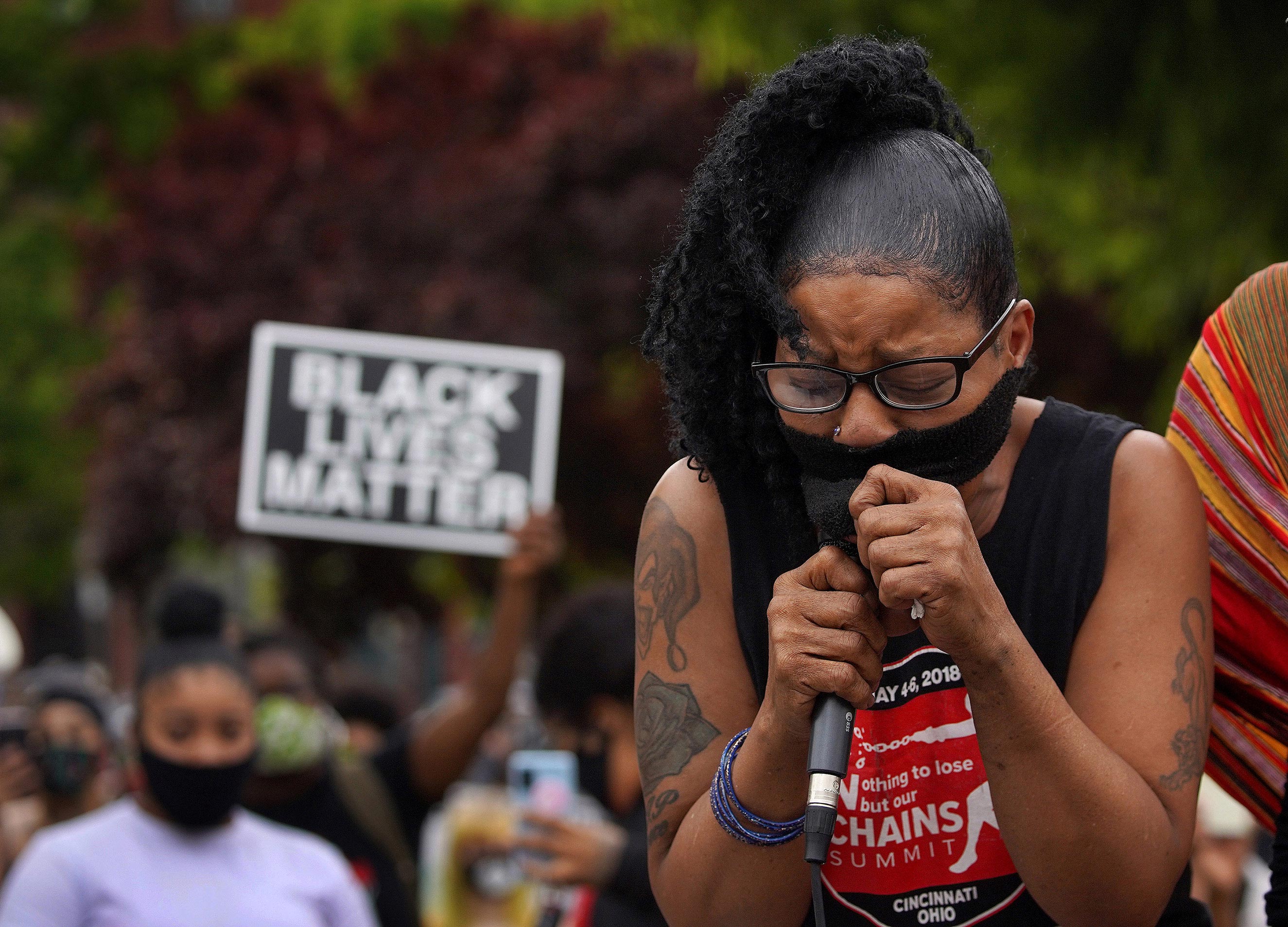 A Black woman wearing a protective face mask tearfully spears into a microphone during a rally to end racism. A person behind her holds up a sign that reads “Black Lives Matter.”