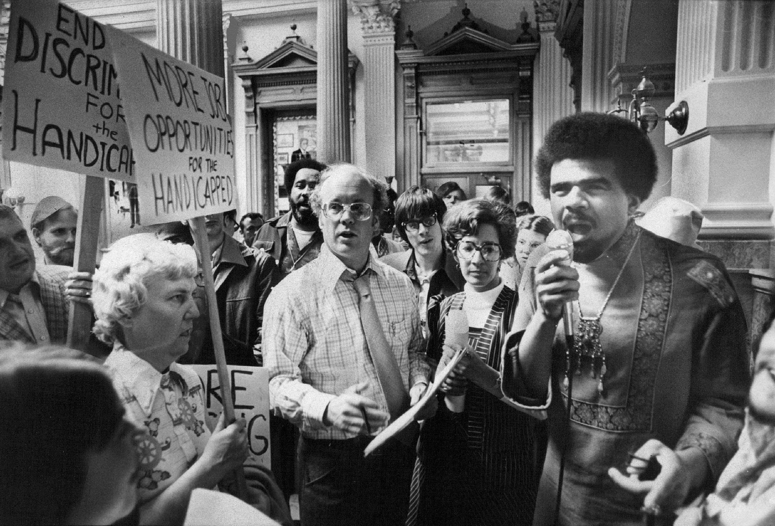 In a vintage photograph, a Black man speaks passionately into a microphone at a rally for disability rights as people around him hold signs demanding equal opportunities and an end to discrimination.