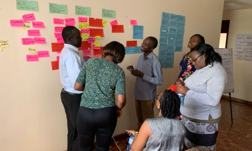 A group of individuals, some standing and some sitting, gather around a wall of brightly colored post-its.