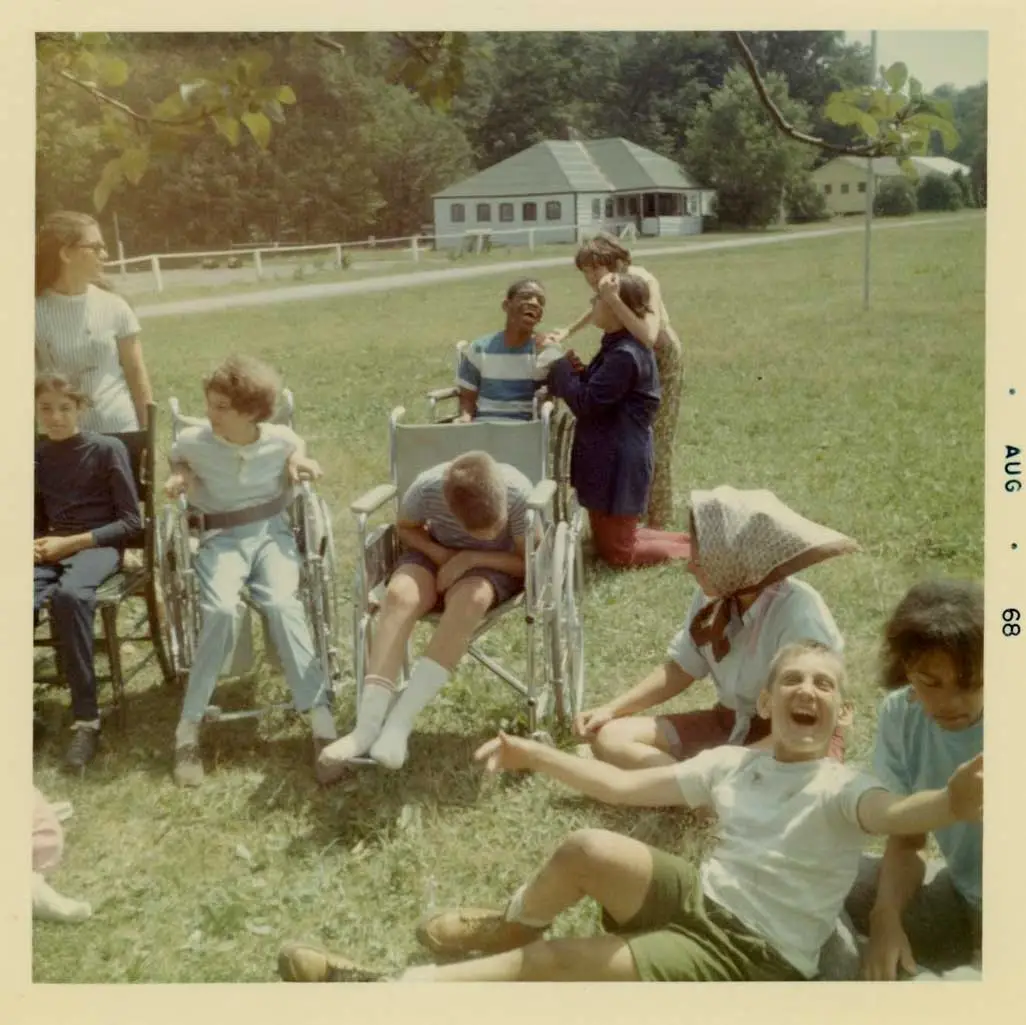 Three boys in wheelchairs laugh on the grass with other young people with disabilities.