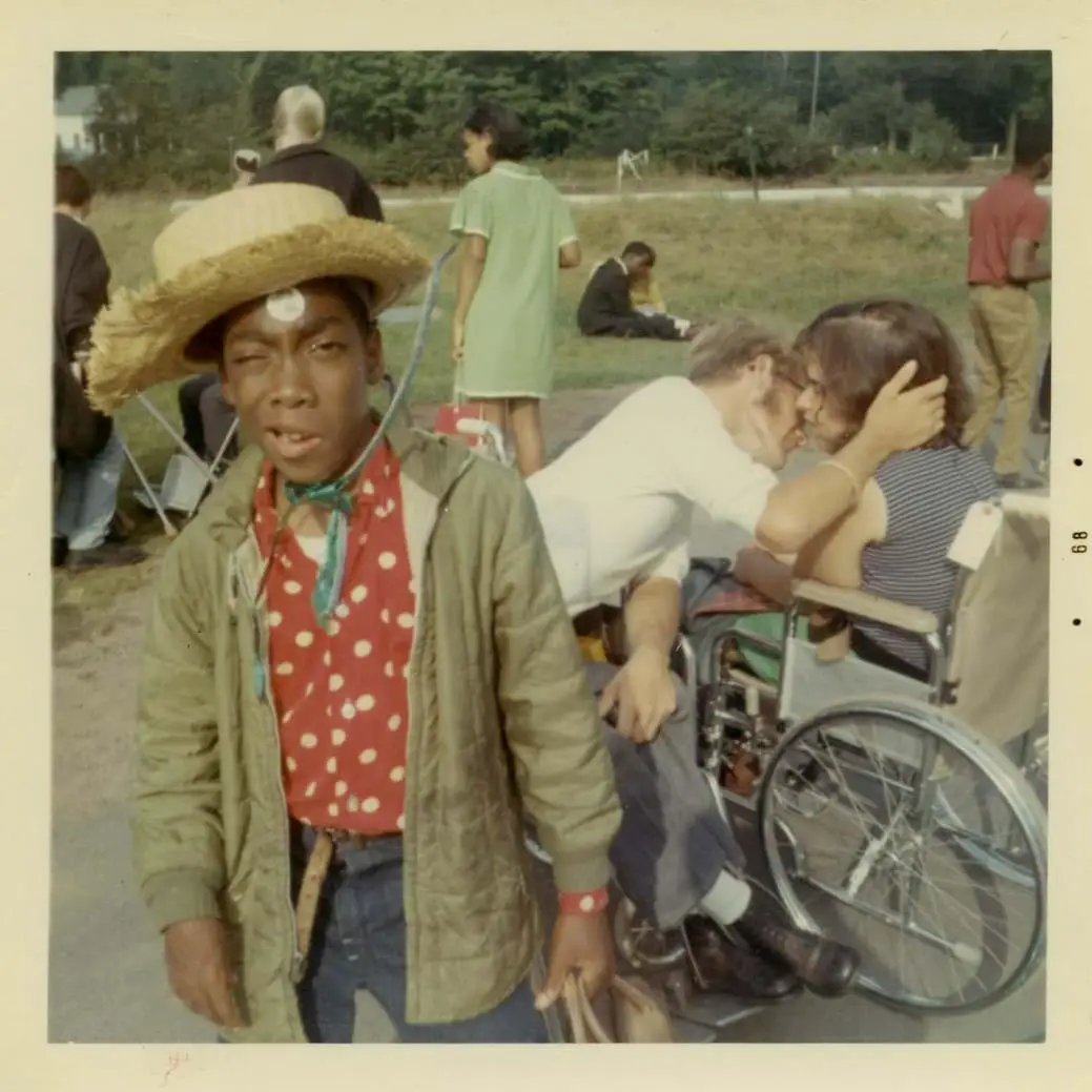 A young boy in a straw hat winks for the camera and two people in wheelchairs share an intimate moment behind him.