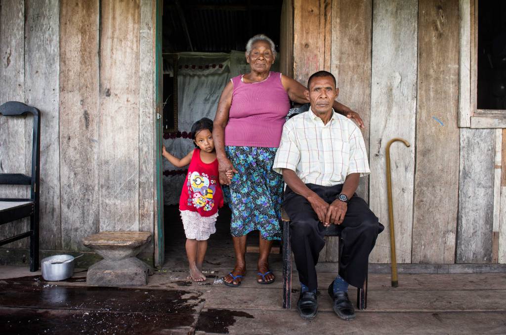 A Honduran man sits in front of a wooden house surrounded by an older woman and a child. There is a cane leaning against the wall behind the man.  Photo: Antonio Busiello