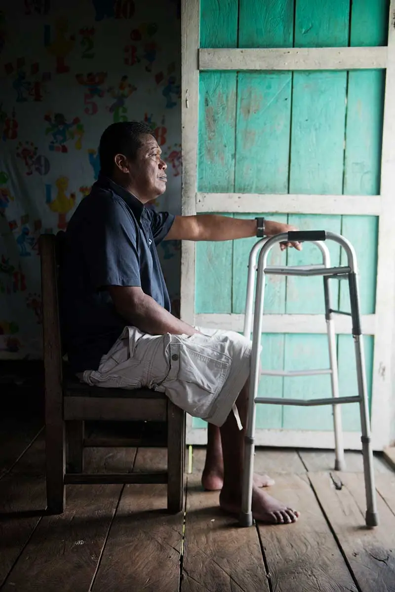  A Honduran man in a blue shirt and white cargo shorts sits on a chair, looking out his front door with his left hand resting on a walker.