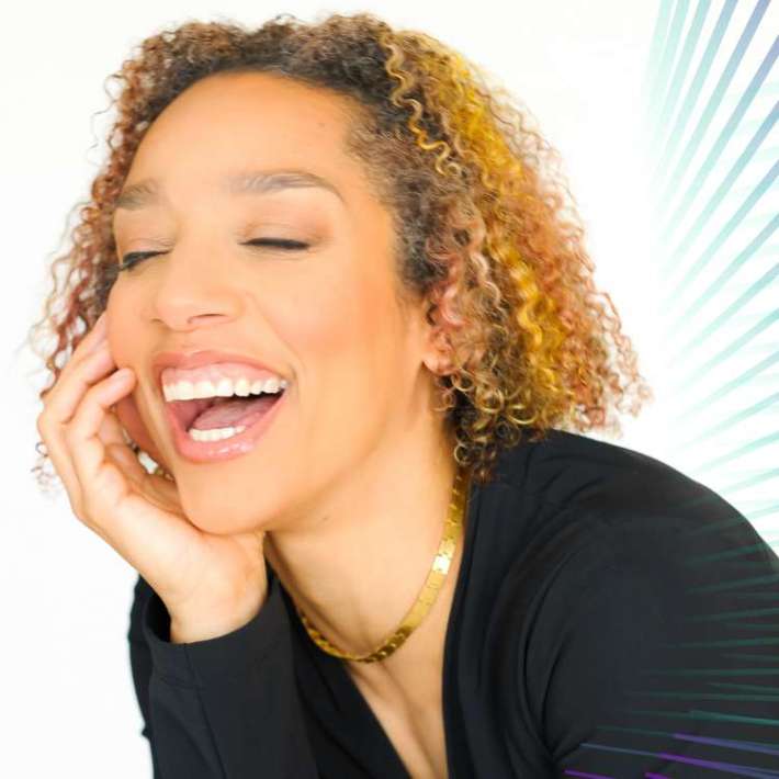 A Black woman leans forward and smiles brightly, teeth showing and eyes closed, as she rests her chin in her palm. She has light brown skin, curly shoulder-length hair with subtle highlights, and wears a black blouse and sleek gold necklace.