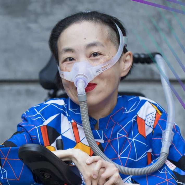 A Chinese-American woman wearing lipstick and a translucent mask over her nose and attached to a tube smiles warmly at the camera. She has cool light skin, short black hair, wears a dark shirt with a geometric pattern, and folds her hands on the control panel of her power chair.