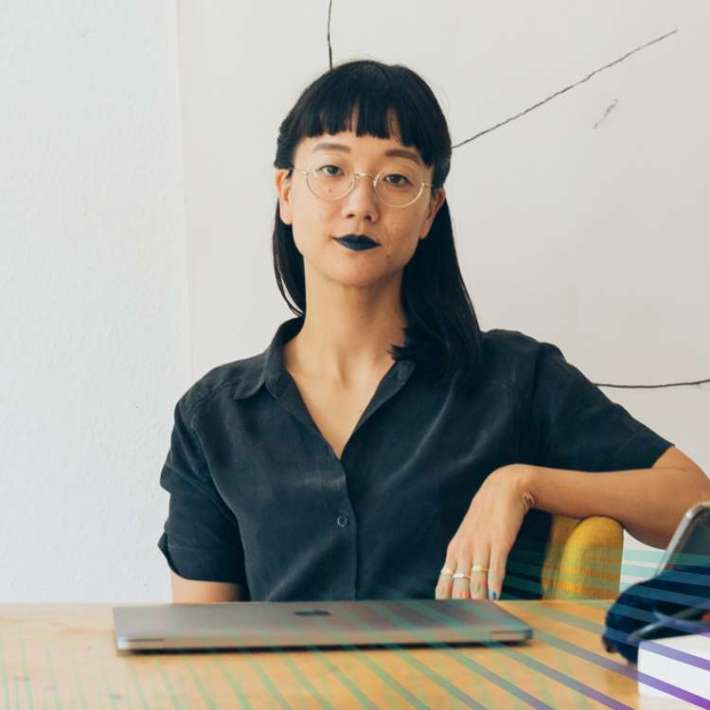 A Korean-American woman sits at her studio desk in a chair and looks coolly at the camera. She has warm light skin, long straight black hair, blunt bangs, and wears dark lipstick, round glasses, a casual black button-down, and long striped shorts.
