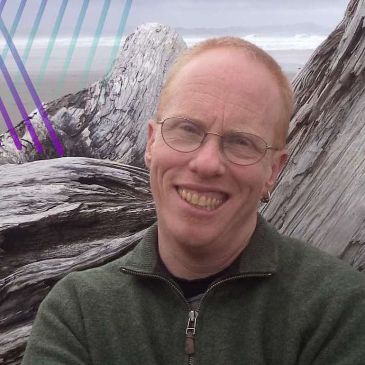 A white nonbinary trans person with a blond buzzcut, oval glasses, and earring poses among wooden logs on a misty beach. He wears an dark fleece and smiles brightly.