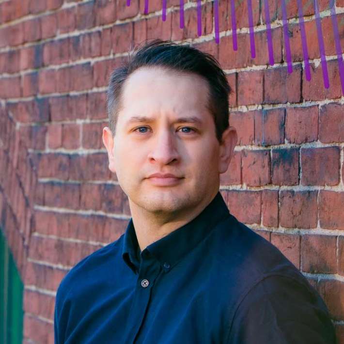 A white-presenting man with short combed dark hair, light skin, and a cleft chin poses in front of a brick wall and stares at the camera with a focused expression. He wears a dark button-down shirt.