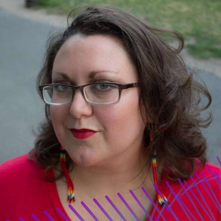 Jen, a Two-Spirit, Cherokee person with warm light skin, shoulder-length dark hair, narrow square glasses, and defined brows, smiles at the camera. Jen wears lipstick, colorful beaded earrings, and a bright top.