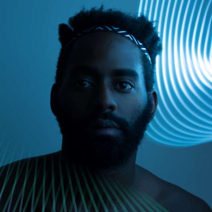 Bathed in light, a shirtless bearded Black man with dark brown skin, a short hi-top fade, and a patterned headband gazes intently into the camera.