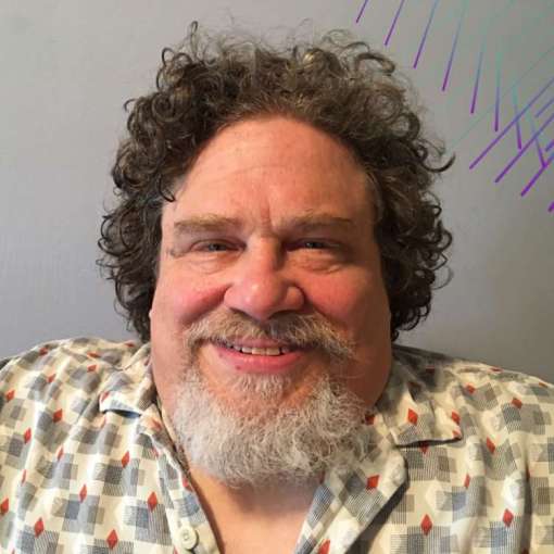 A white man smiles warmly at the camera. He has a grayish-white goatee and mustache, curly dark gray hair, and wears a light-colored geometric patterned button-down.