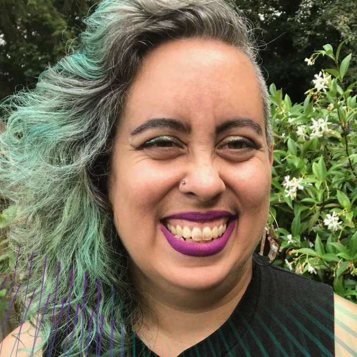 Wearing a sleeveless black t-shirt with a shark graphic, a Sri Lankan, Roma and Irish nonbinary femme with tan skin, curly shoulder-length hair, lipstick, and tattooed arms, smiles brightly at the camera in front of a blooming jasmine vine.