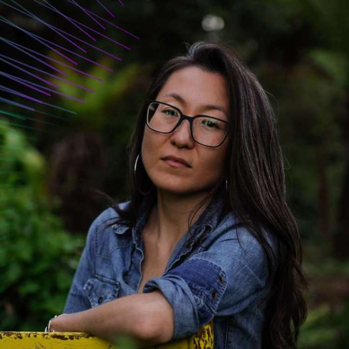 A Korean woman with warm light skin, long dark hair, big square glasses, and wearing a denim shirt sits casually outside in a wooden chair backdropped by foliage and stares confidently into the camera.
