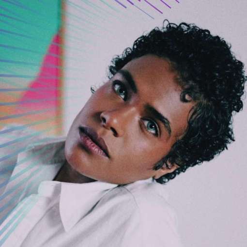 Leaning back casually with a serene expression, a Black nonbinary trans person with medium brown skin, and short curly black hair gazes into the camera. He has a defined brow and wears an unbuttoned white shirt.