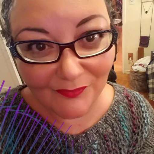 A Japanese-Haitian nonbinary person with tan skin, narrow square glasses, lipstick, and arched brows smiles at the camera. They pose in a grey knit sweater.