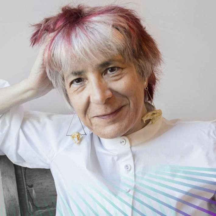 A white woman with short white hair with subtle lowlights, geometric earrings, and a crisp white collarless shirt poses seated, resting one arm on the back of her chair and smiling warmly.