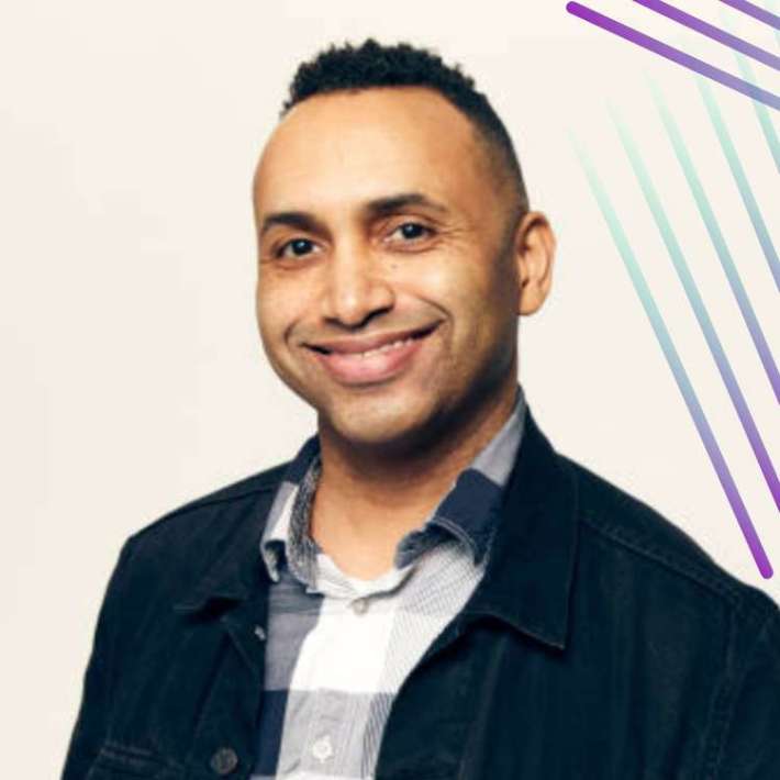 A Black man with warm medium brown skin, hair faded on the sides and back with twists on top, is wearing a plaid shirt with a black jeans jacket against a white background. He smiles enthusiastically at the camera.