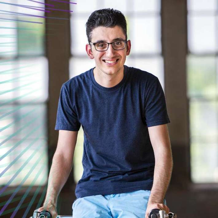 A man of Lebanese descent with light warm skin and short black hair is wearing square glasses, a t-shirt, shorts, and lower leg braces. He poses with his metallic walking frame and smiles brightly at the camera.
