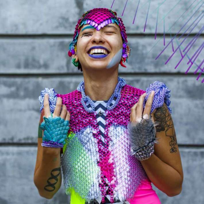 A Filipinx nonbinary person throws back their head and smiles joyously. They have tan skin and wear a reflective headpiece made of metallic scales. They're wearing a radiant chain link top with protruding sculptural shoulders, colorful gloves with metallic scales, and dark lipstick.