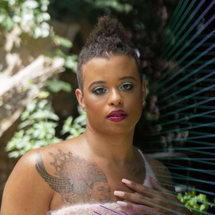 A Black nonbinary trans person with a high bun of dark brown hair, eye shadow, and dark lipstick poses in a wooded area. She looks softly into the camera with one hand placed on her chest and wears a one-shoulder white top that showcases her tattoos.