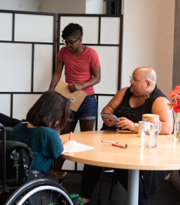 A person in a red shirt stands holding a clipboard behind a round table. At the table, another person writes on paper, and a third person with a shaved head looks at a tablet. A vase of red flowers is on the table.