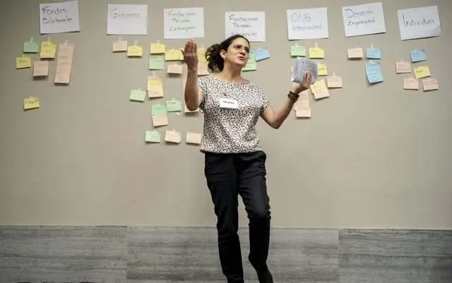  A woman wearing a patterned blouse and black pants stands in front of a wall of sticky notes with her arms open.