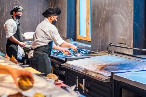 Two chefs wearing black hats and face masks work in a modern kitchen. One chef tends to a pan on a stove, while another stands further back. A large grill area is prominent in the foreground.