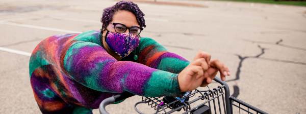 Sami Schalk wearing a bright colored sparkling bodysuit, eyeglasses, and an iridescent sequin face mask leaning over a supermarket shopping cart