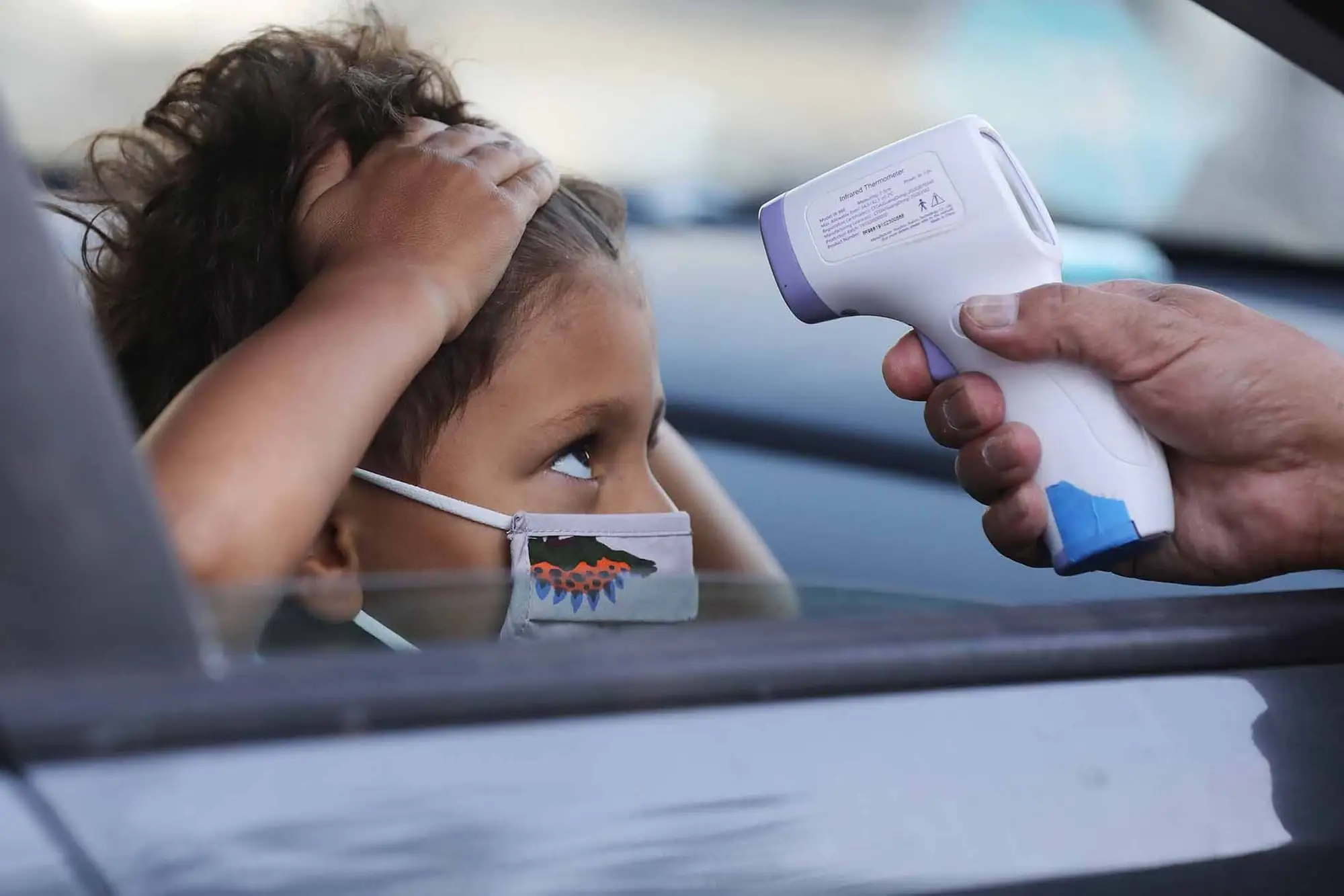 A young student inside a car wearing a medical mask pulls their hair back as a hand with a thermometer is pointed to their head.