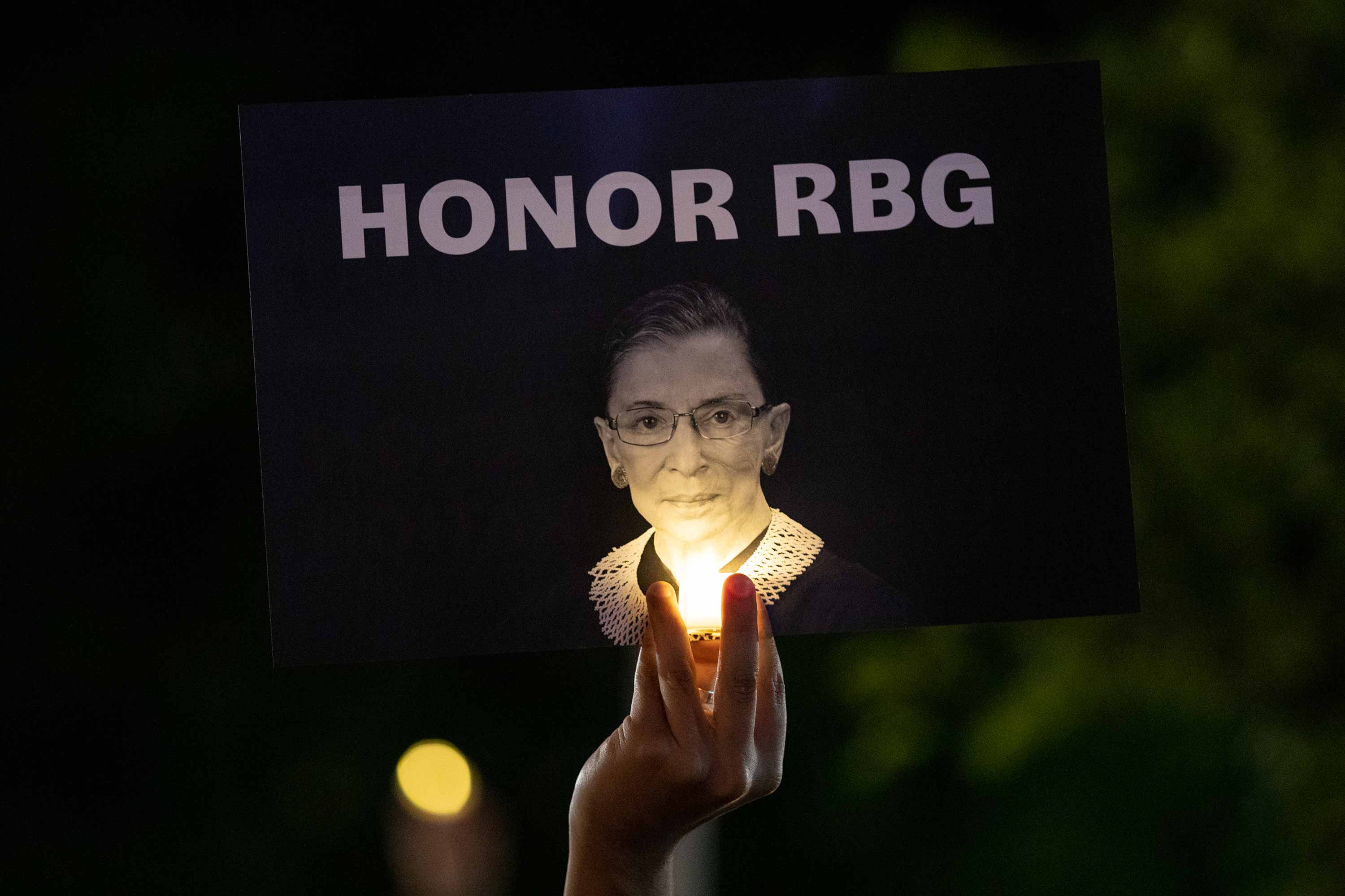 A hand holds up a sign that reads “Honor RBG” while lit by a small handheld light. 