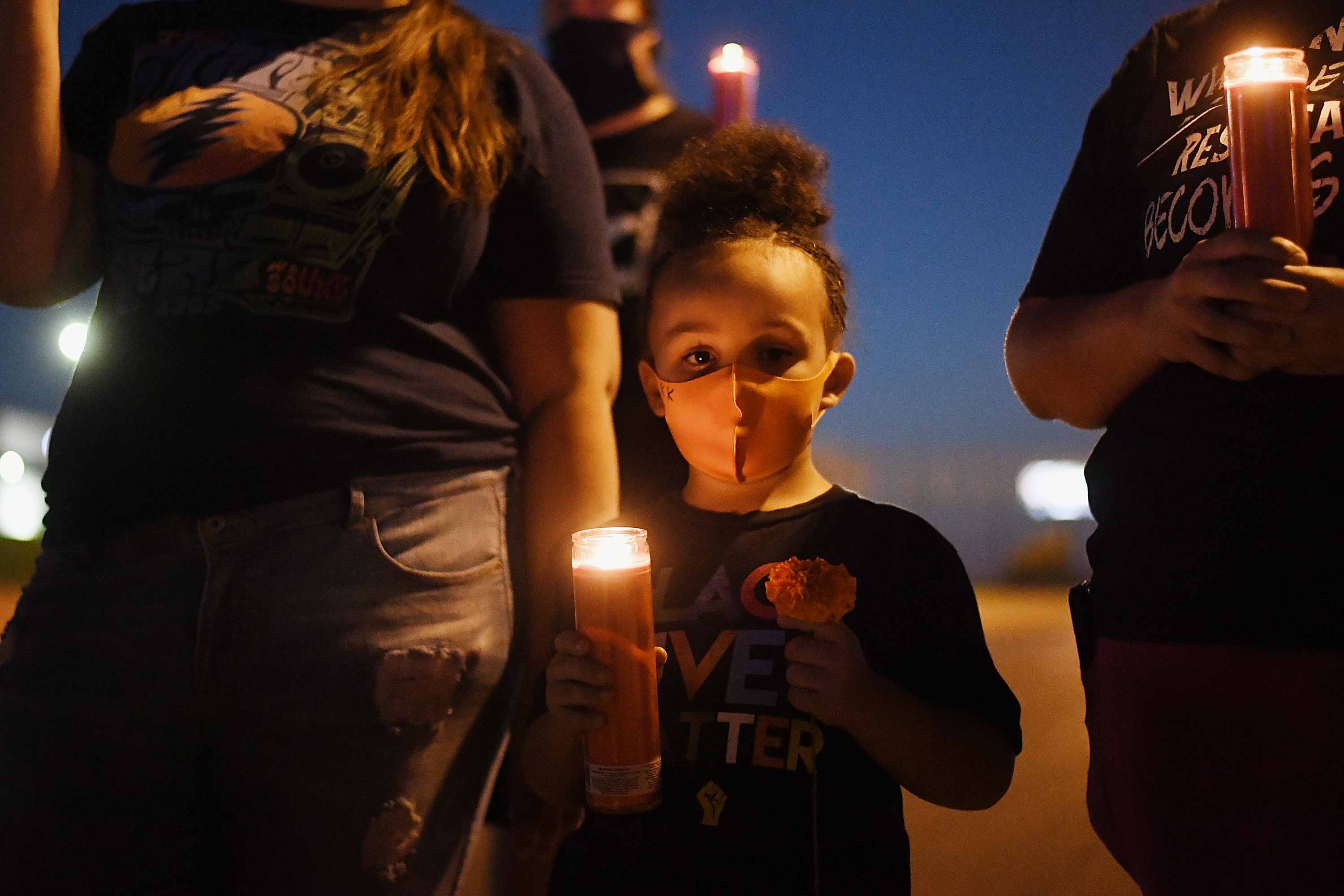 A young Black girl wearing a protective mask holds a candle in her hand while surrounded by adults.