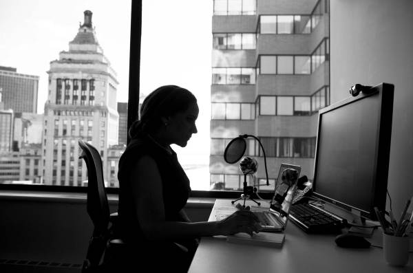 Black and white photo of a woman working in an office, at a computer. City buildings can be seen in the background, through her windows. Photo by #WOCinTech