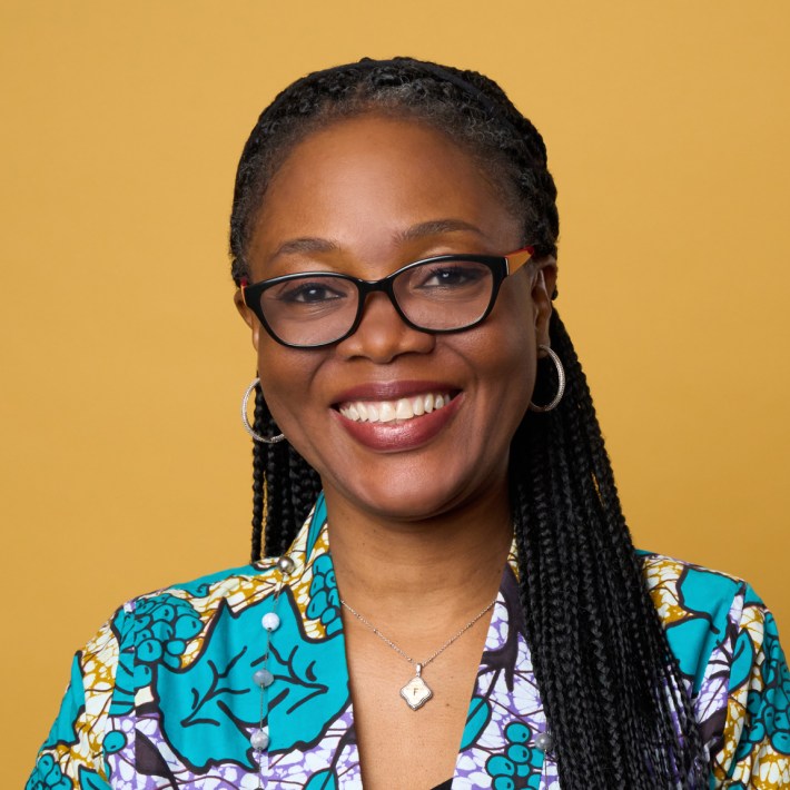 Portrait Olufunke Baruwa wearing a red top and black frame glasses against a mustard yellow background.