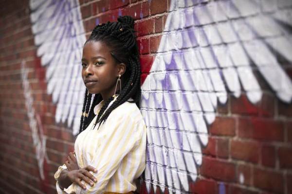 Amanda Gorman is wearing a yellow and white striped blouse, standing in front of a brick wall with angel wings painted on it. Photo Peter Stevens / Flickr