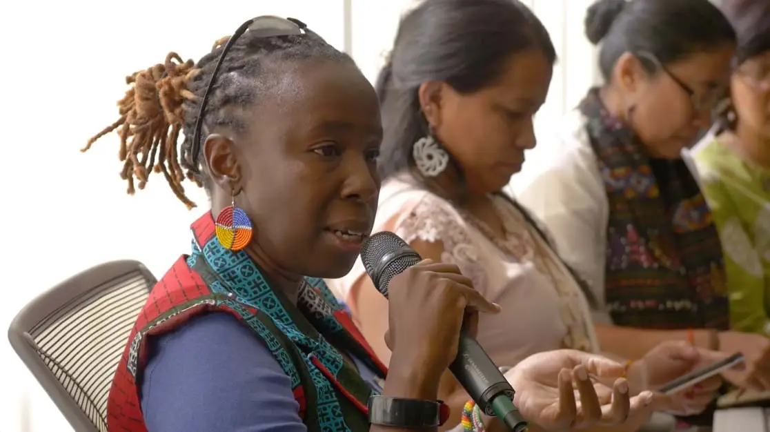 A seated woman with braided hair and multi-colored bead earrings and vest, holding a microphone and gesturing with her hand with other women seated beside her on their phones.