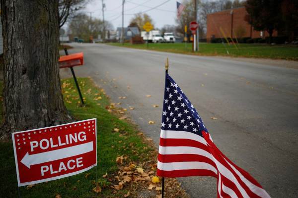 A red sign on the left reads "Polling Place" with a white arrow while an American flag flows in the wind in the center on a quiet neighborhood street. 