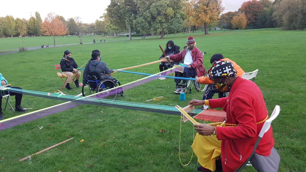 People in colorful clothing and face masks weave multicolored yarn onto a geometric structure outside at a spacious green park