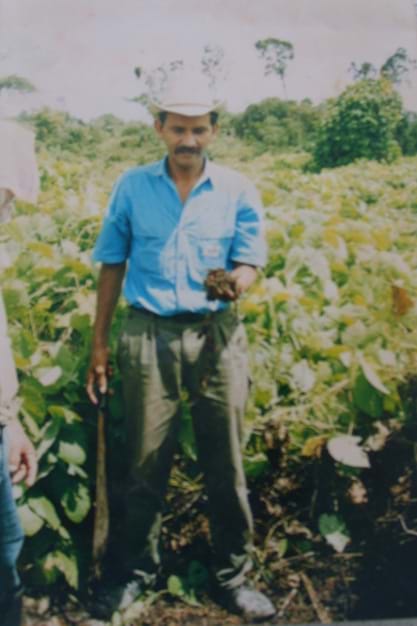 Marcedonio Cortave wears a brimmed hat, blue button up shirt, and olive green pants while standing in a field and holding a handful of soil in his hand.