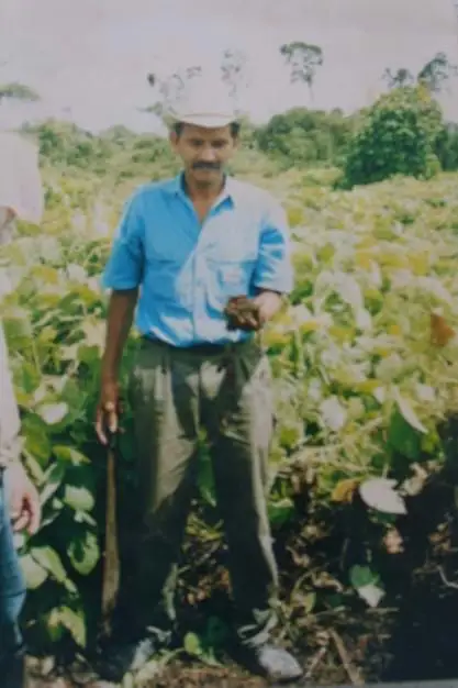 Marcedonio Cortave wears a brimmed hat, blue button up shirt, and olive green pants while standing in a field and holding a handful of soil in his hand.