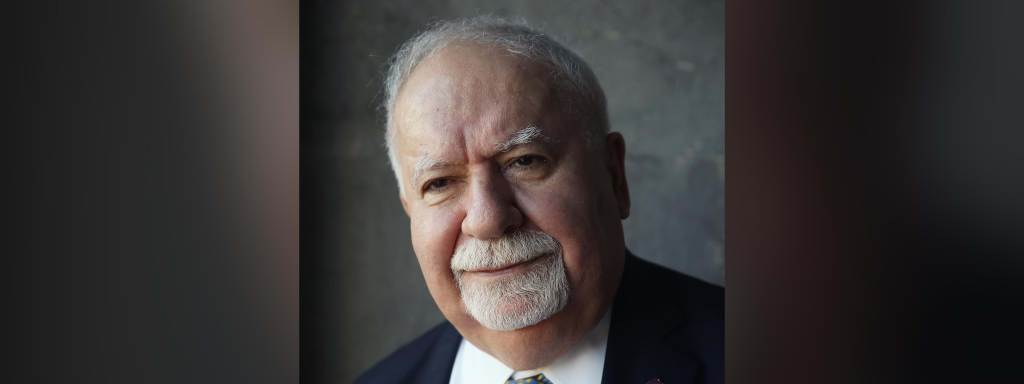Vartan Gregorian wearing a navy suit, white shirt, and polka-dotted tie smiles against a gray backdrop