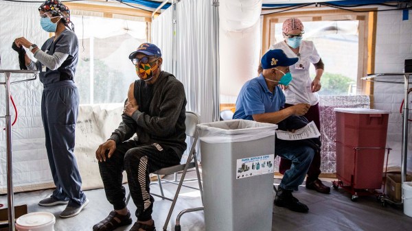 Four individuals wearing masks are in a COVID-19 vaccination center. Two of the individuals are student nurses, while the other two are there to receive their COVID-19 vaccinations.