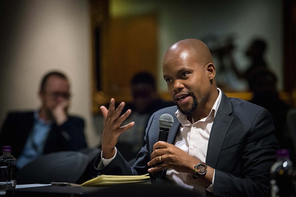 Mbongiseni Buthelezi, a Black man with a beard and shaved head, wears a white checkered dress shirt  and navy blazer, holding a microphone speaking in a conference room.